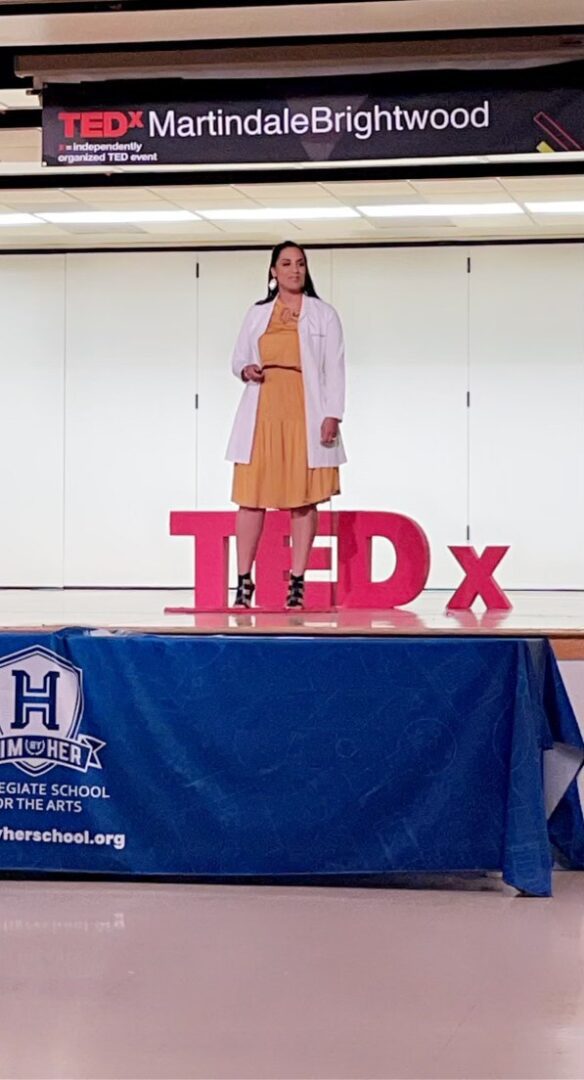 Woman speaking on stage at a TEDx event wearing a yellow dress and white cardigan.